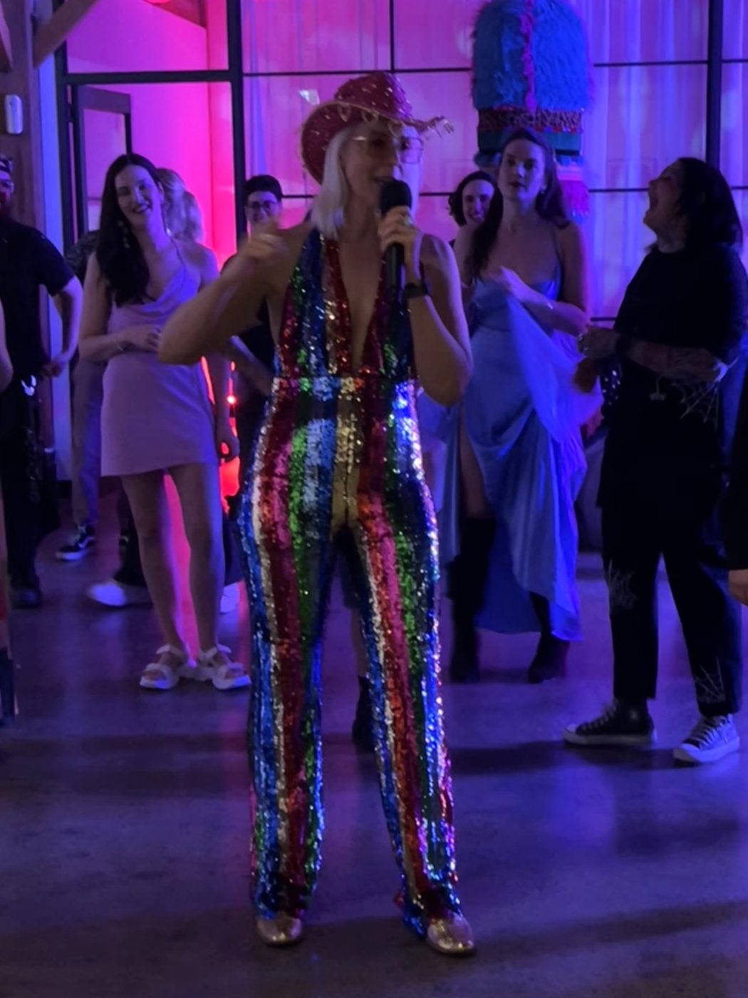 Kirsten Mann in a rainbow sequin jumpsuit hosting on the mic